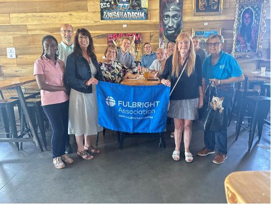 A group of people sitting and standing around a restaurant table holding a banner for the Fulbright Association NC Chapter.